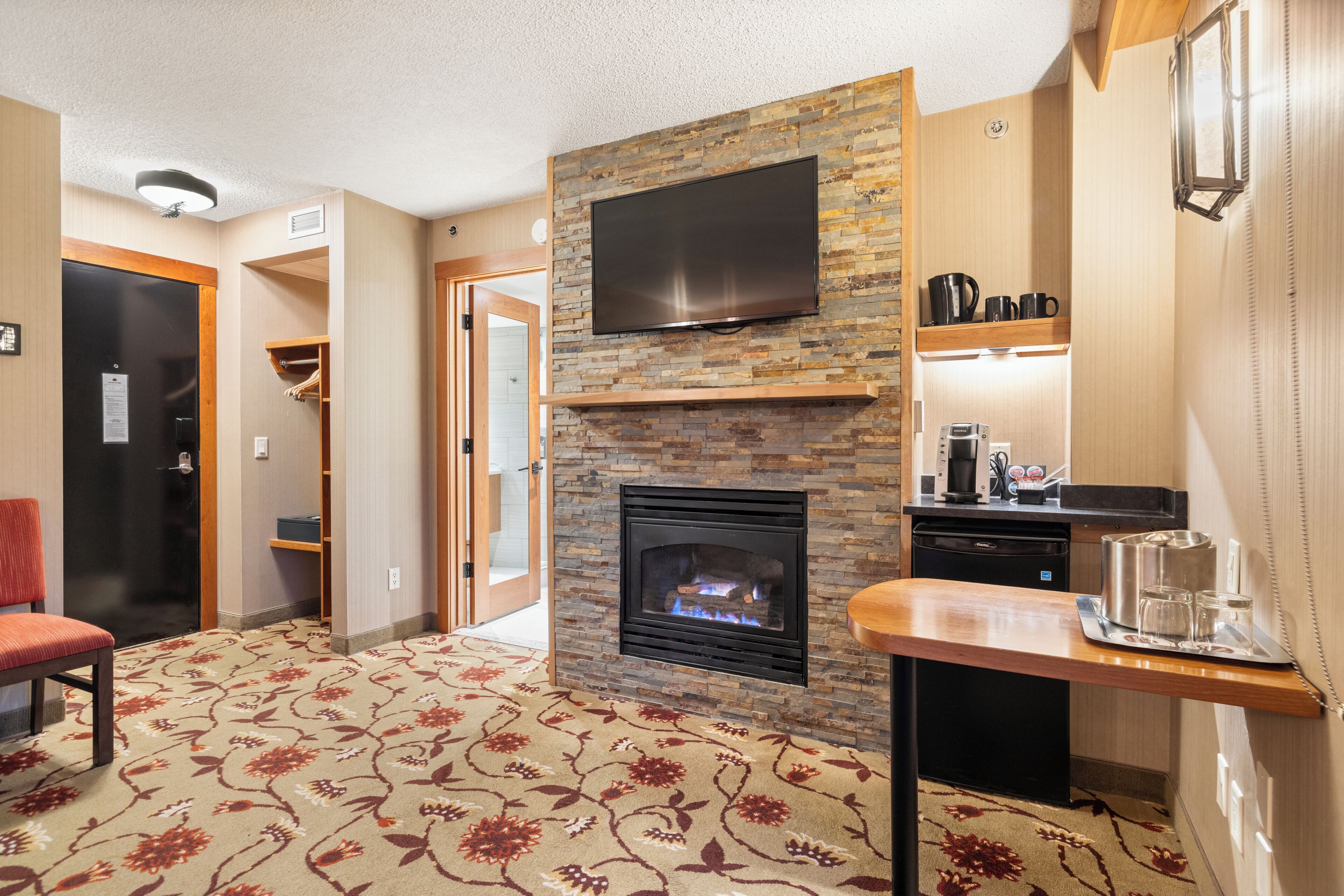 Habitación de hotel con chimenea, televisor de pantalla plana sobre ella, pequeña cocina con cafetera y alfombra estampada.