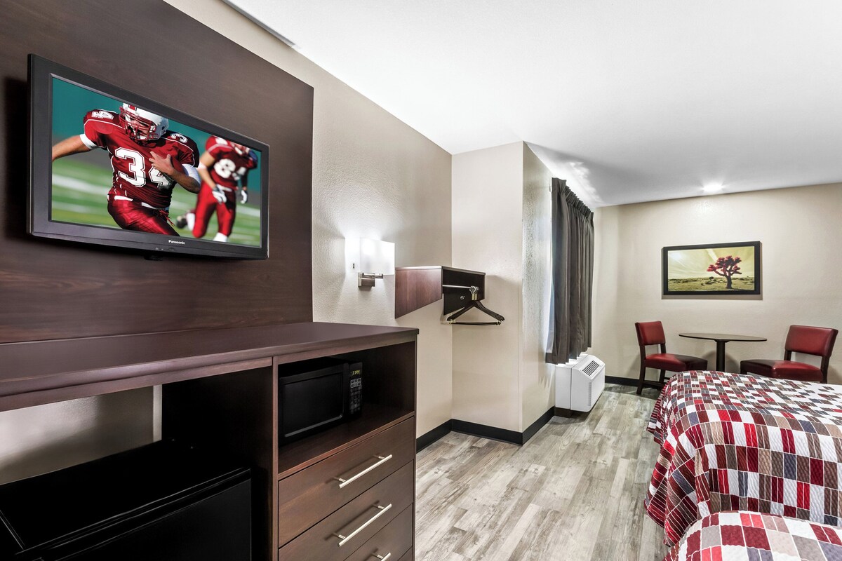 Une chambre d’hôtel avec un téléviseur fixé au mur, un lit recouvert d’une courtepointe à carreaux, un bureau avec un ordinateur, une chaise, une petite table et un tableau accroché au mur.