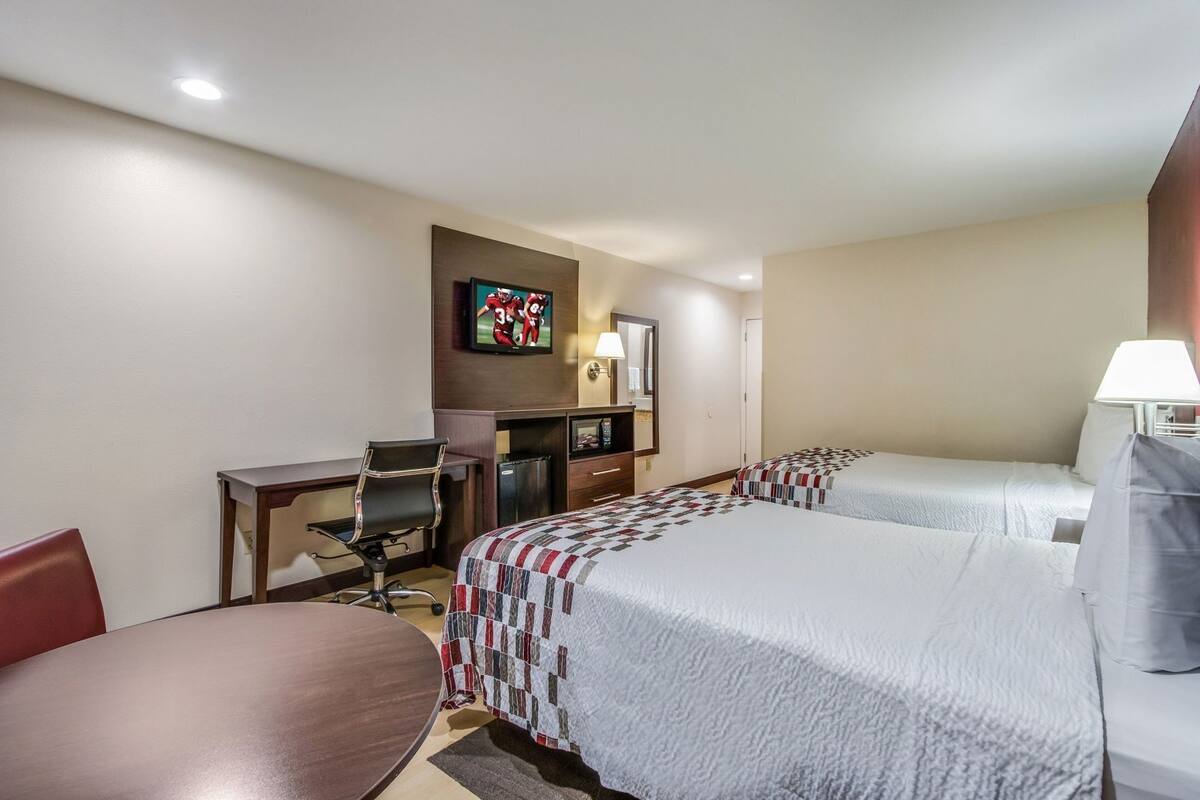 A hotel room with two beds, a desk, a chair, a TV, and a lamp.
