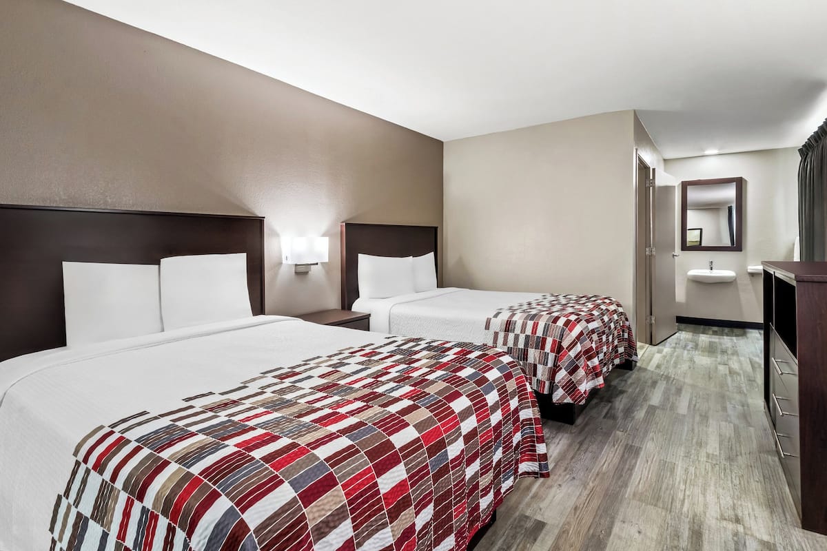 A hotel room with two beds, a nightstand, a lamp, a mirror, and a wooden floor.