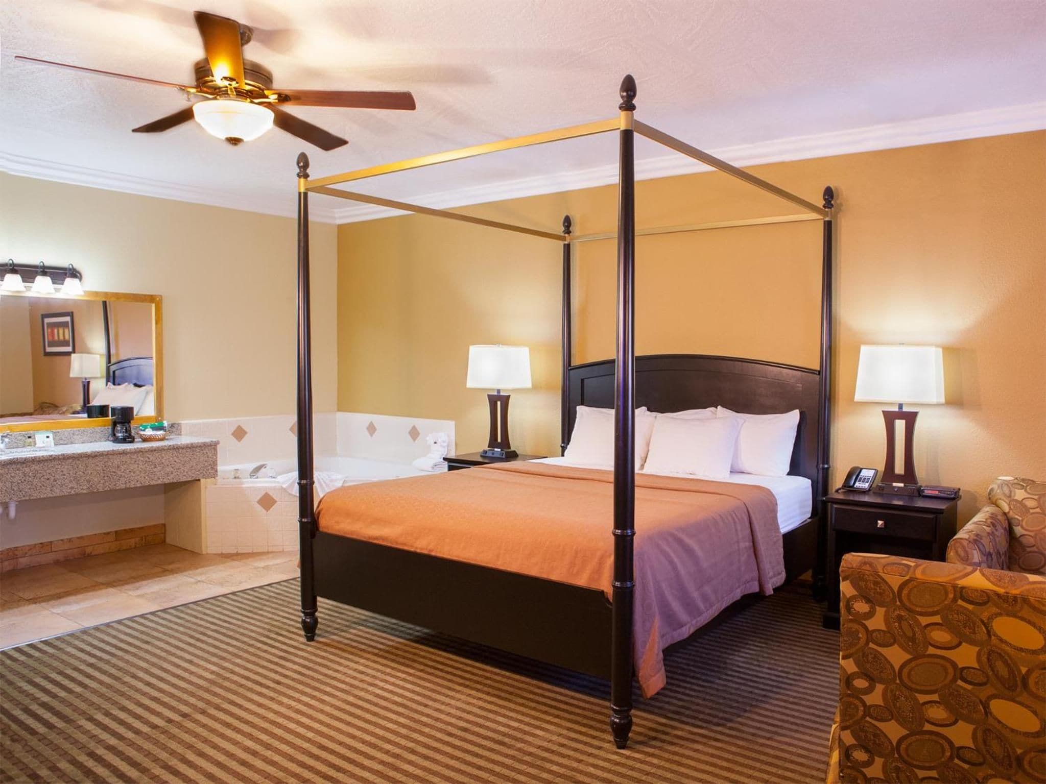 A hotel room with a four-poster bed, a sitting area with a chair, a vanity with a mirror, and a ceiling fan.