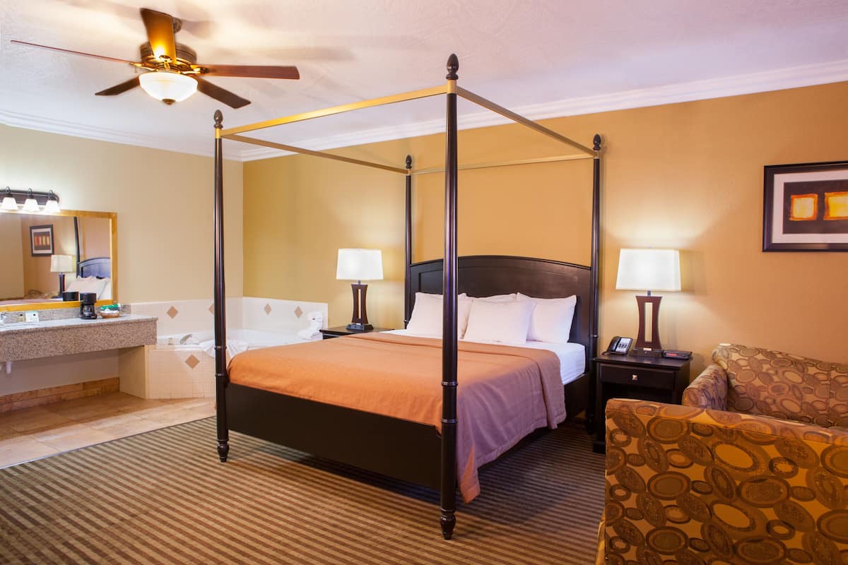 A four-poster bed with a headboard, two bedside tables with lamps, a chair, and a bathroom with a sink and mirror.