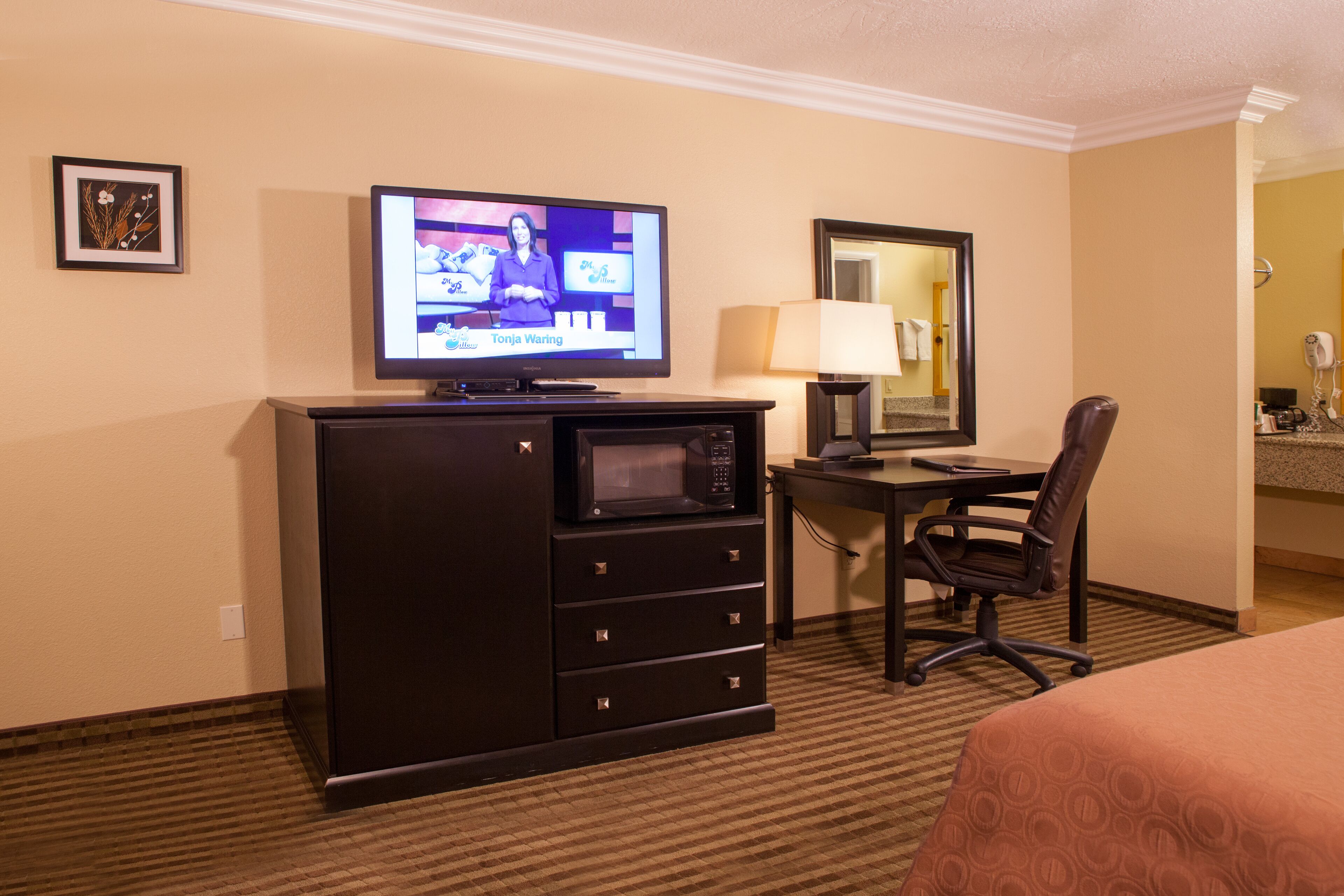 Habitación de hotel con cama, escritorio con computadora, microondas, silla, lámpara y televisor mostrando un noticiero.
