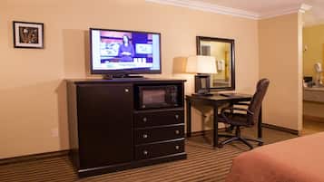 Habitación de hotel con cama, escritorio con computadora, microondas, silla, lámpara y televisor mostrando un noticiero.