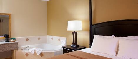 Habitación de hotel con una cama grande, baño con bañera, mesita de noche con lámpara y un espejo en la pared.
