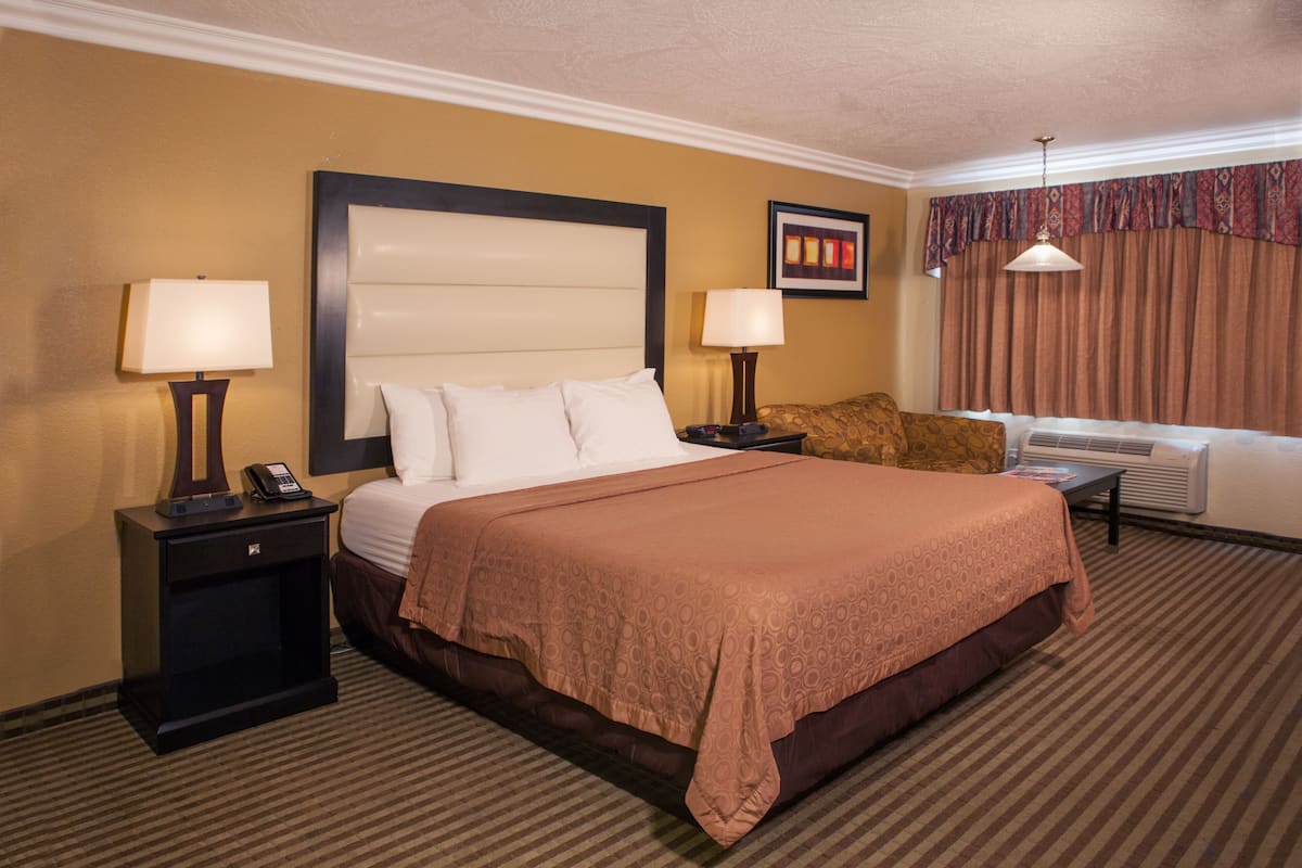 A hotel room with a large bed, two bedside tables with lamps, a nightstand, a wall-mounted telephone, a framed picture, a window with curtains, and an air conditioning unit.