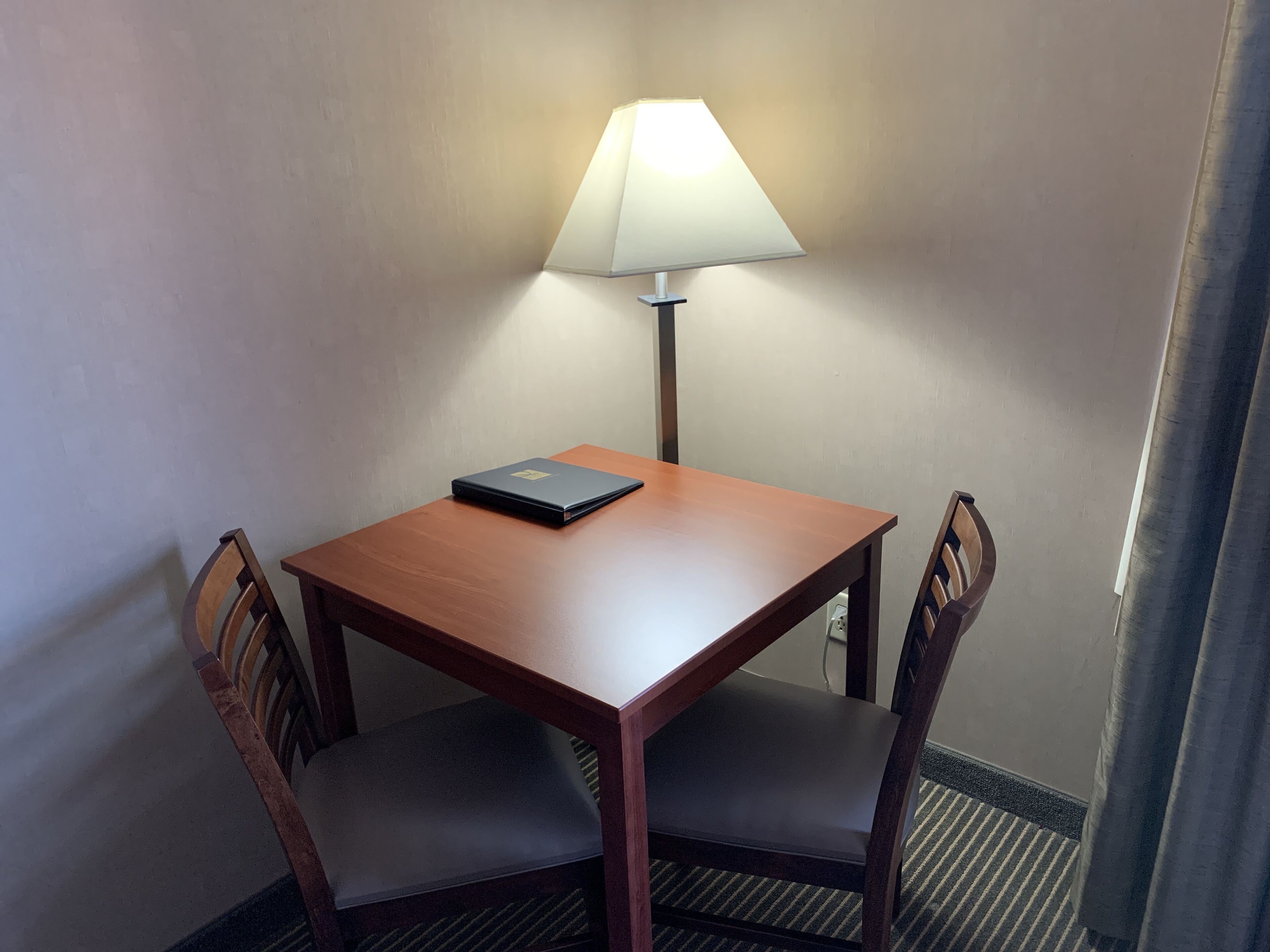 A small wooden table with a laptop and a lamp, two chairs, and a plain wall.