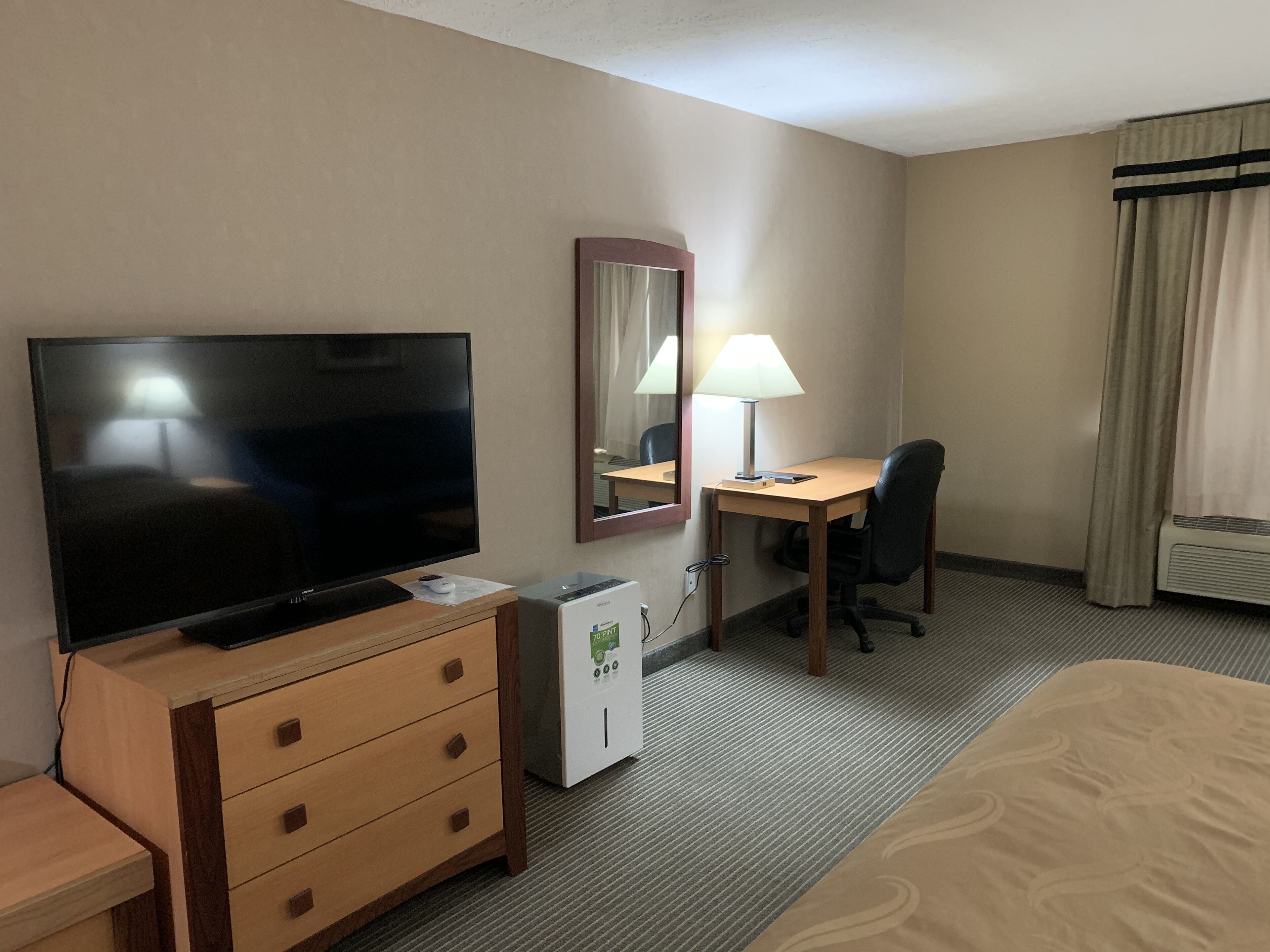 Una habitación de hotel con televisión sobre un mueble de madera, un escritorio con lámpara, un espejo y una cama con edredón estampado.