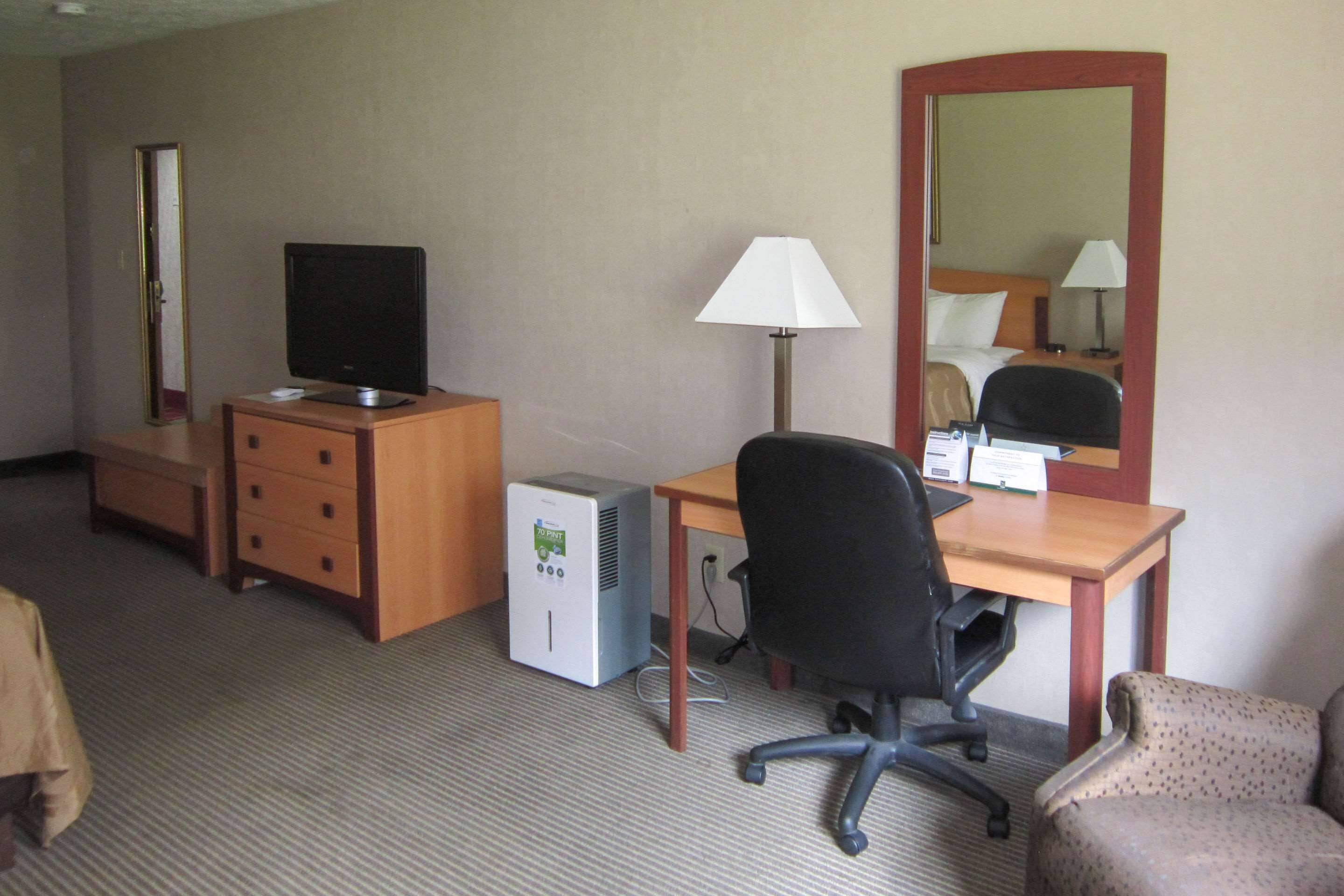 Hotellihuone, jossa on puinen työpöytä, musta toimistotuoli, pieni pöytälamppu, televisio, minijääkaappi, peili ja kuviollinen nojatuoli.