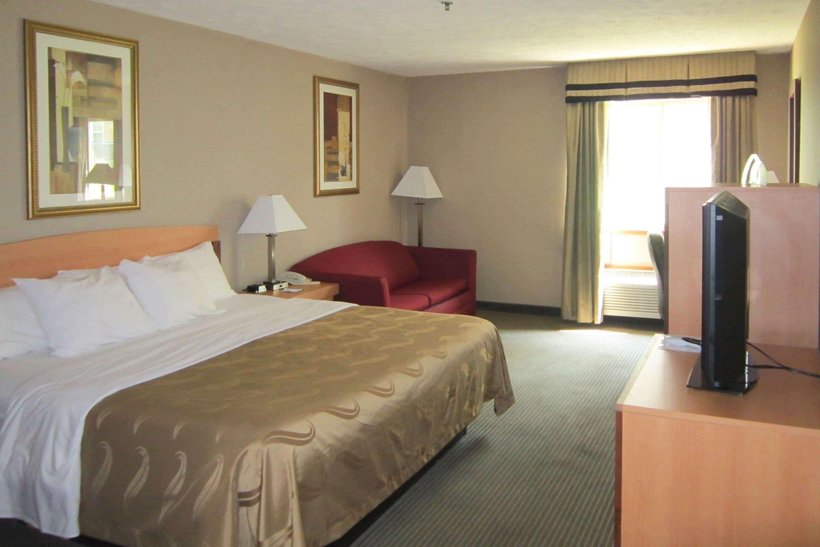 Een hotelkamer met een groot bed, een bureau met een televisie, een rode stoel, twee lampen en ingelijste foto's aan de muur.