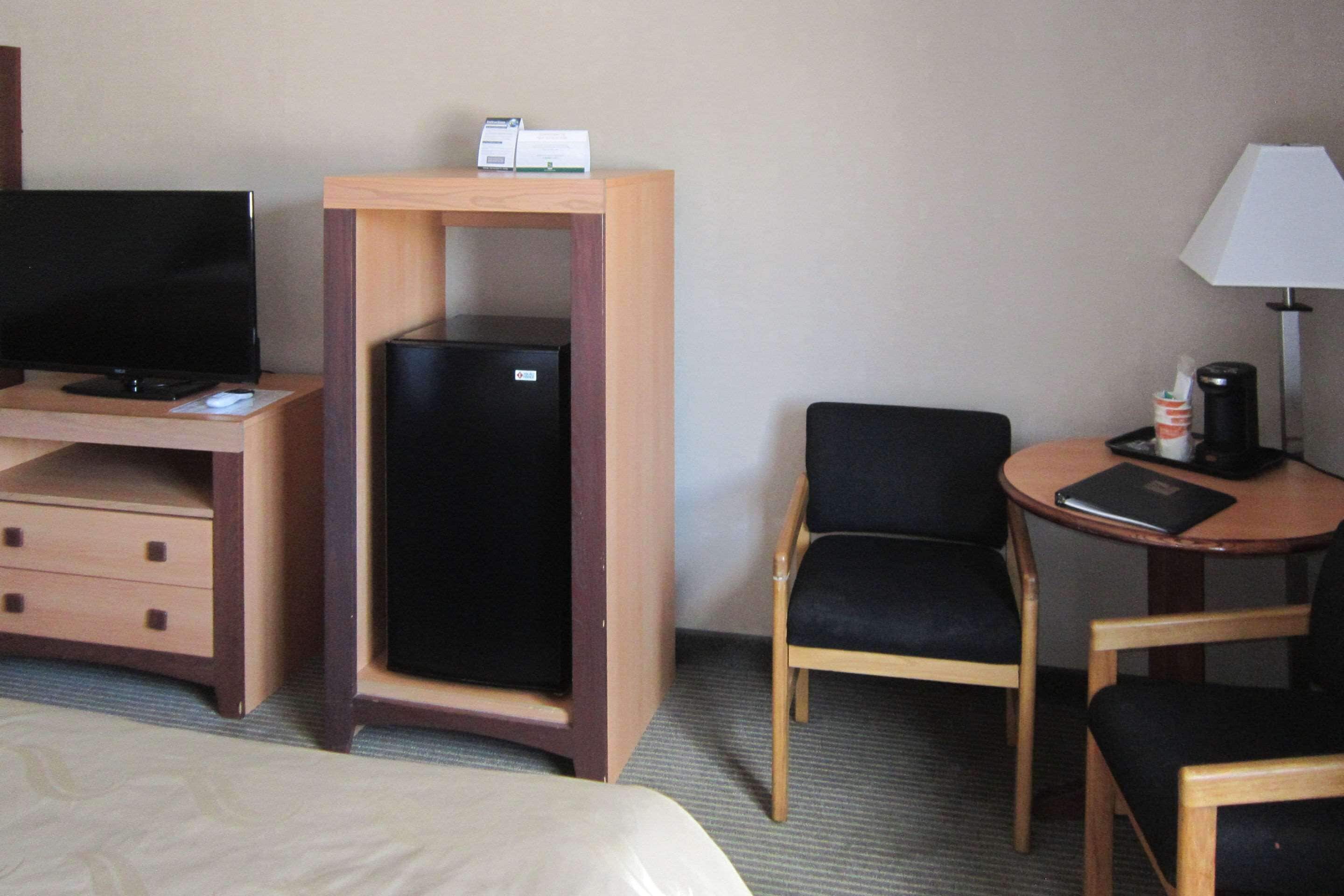 Habitació d'hotel amb llit, escriptori amb ordinador, nevera, cadira, làmpada i cafetera.