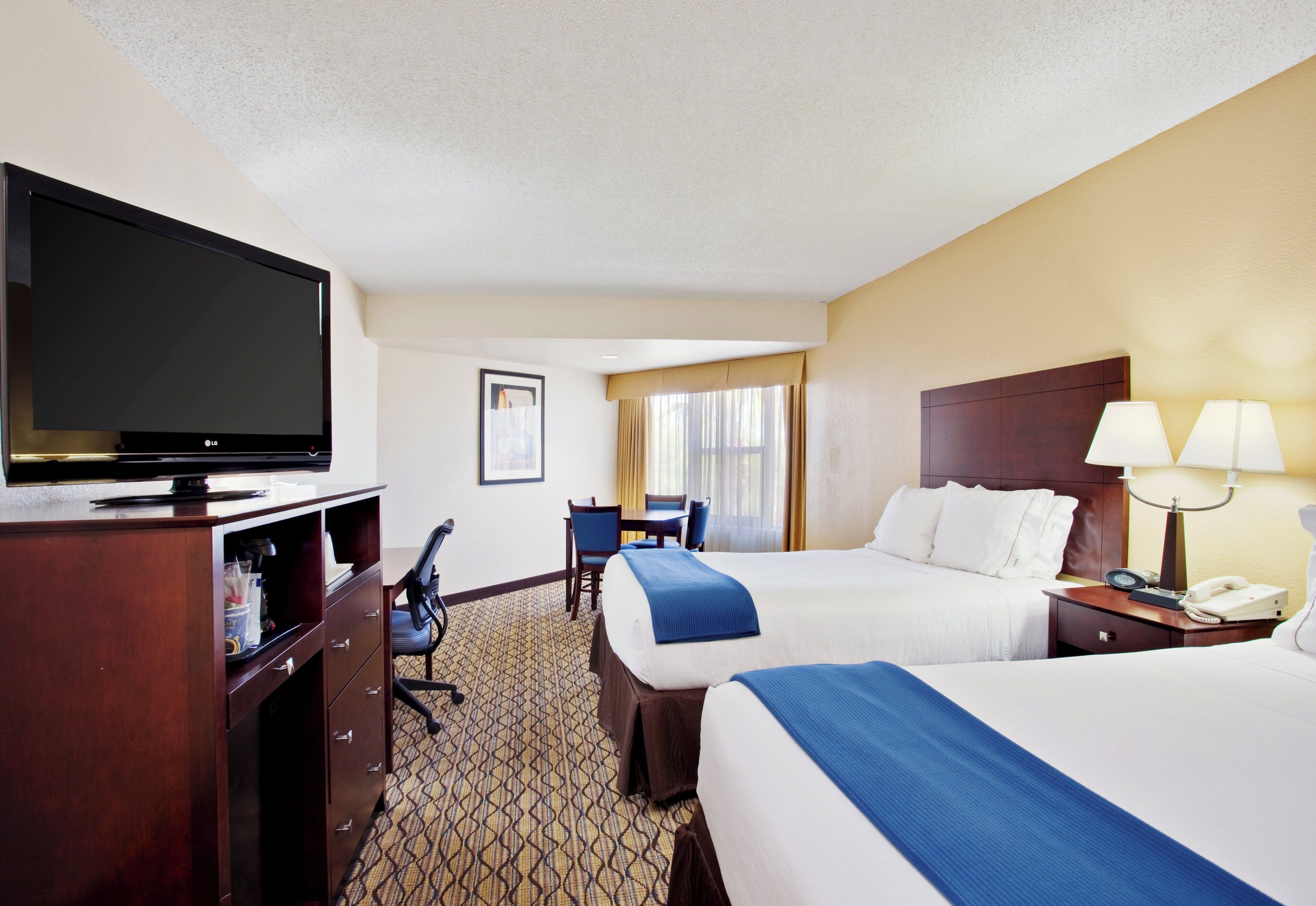 A hotel room with a large flat-screen TV, a desk with a chair, a dining table with chairs, a bed with a blue blanket, and a bedside table with a lamp and a phone.