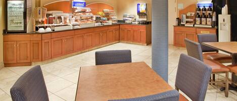 A hotel breakfast area with a self-serve station, seating, and a trash bin.