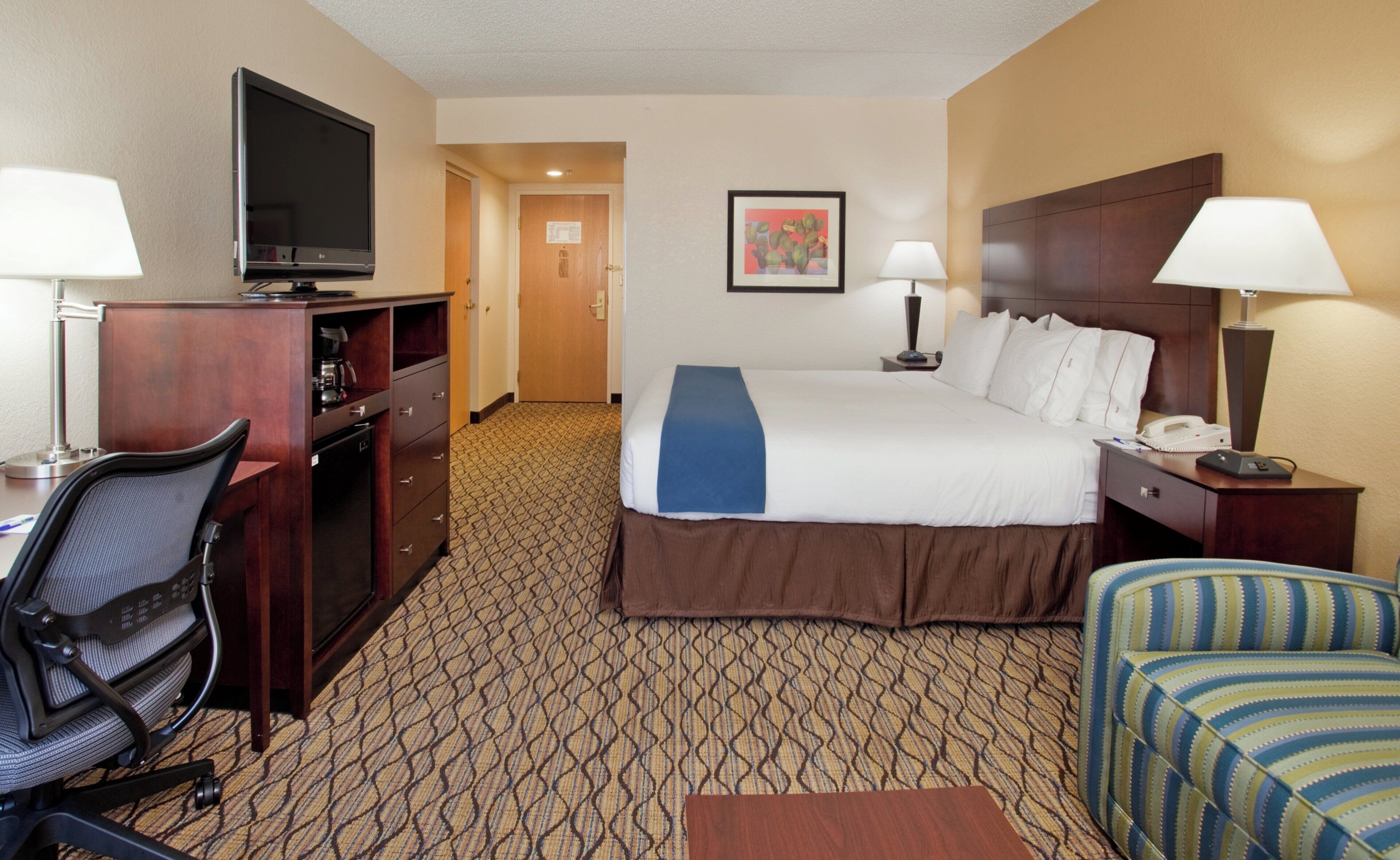 A hotel room with a bed, desk, chair, television, and artwork on the wall.