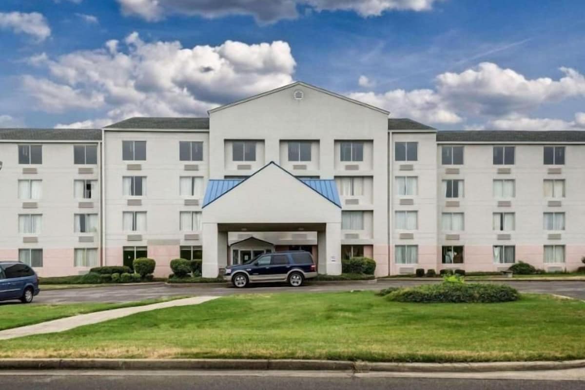 A multi-story hotel with a central entrance, parking area, and a well-maintained lawn.