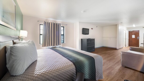 A hotel room with a bed, a desk, a chair, a flat-screen TV, and a wooden floor. - HomeTowne Studios by Red Roof St. Louis - Airport/ N Lindbergh (Hazelwood)