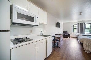 A compact kitchen with a microwave, stove, and sink, a small dining table with chairs, a sofa, and a TV mounted on the wall. - HomeTowne Studios by Red Roof St. Louis - Airport/ N Lindbergh (Hazelwood)