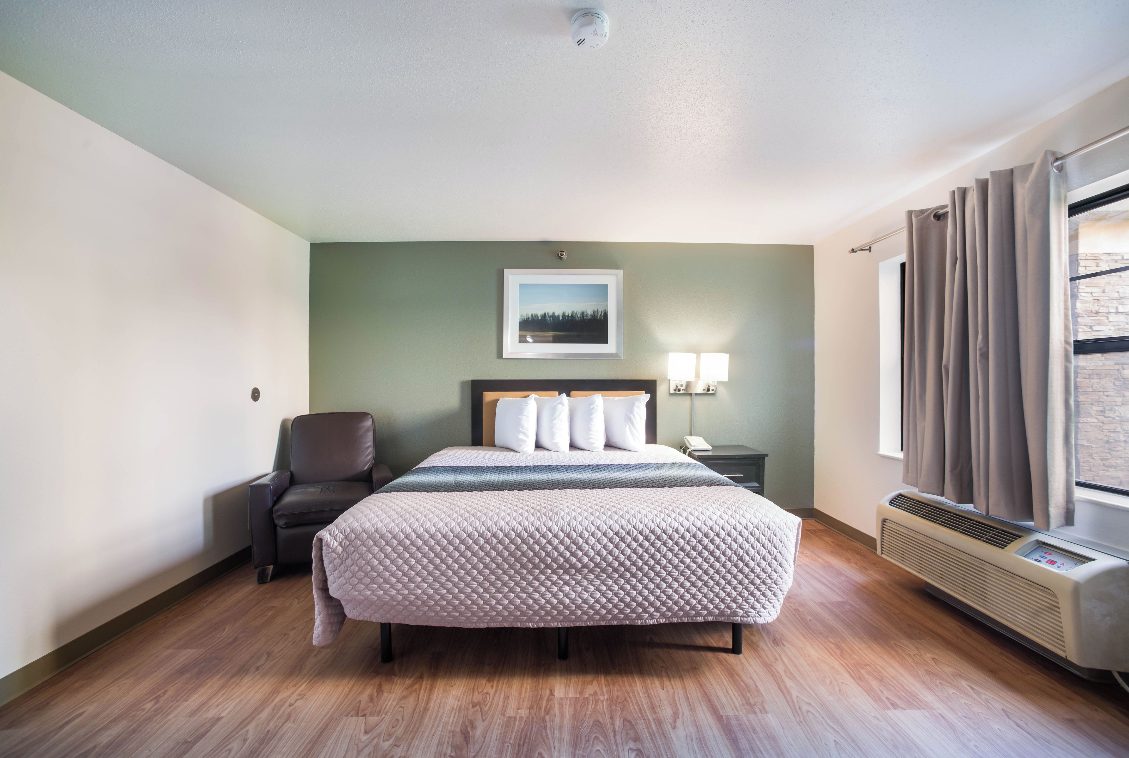 A hotel room with a bed, a chair, a nightstand with a lamp, a picture on the wall, and a window with curtains.
