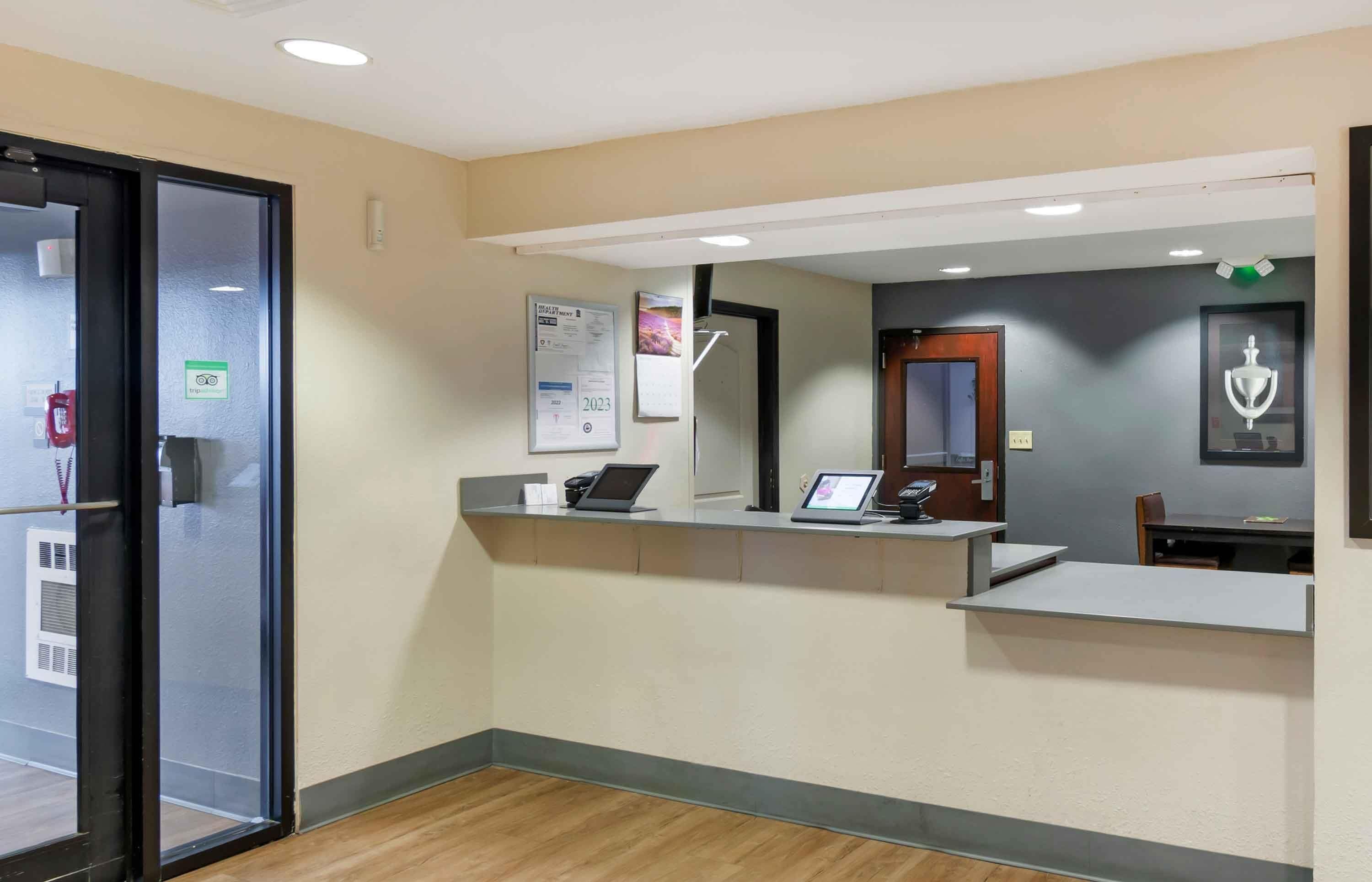 Un área de recepción con entrada por puerta de vidrio, un mostrador de recepción con computadora, un calendario colgado en la pared y un objeto enmarcado.