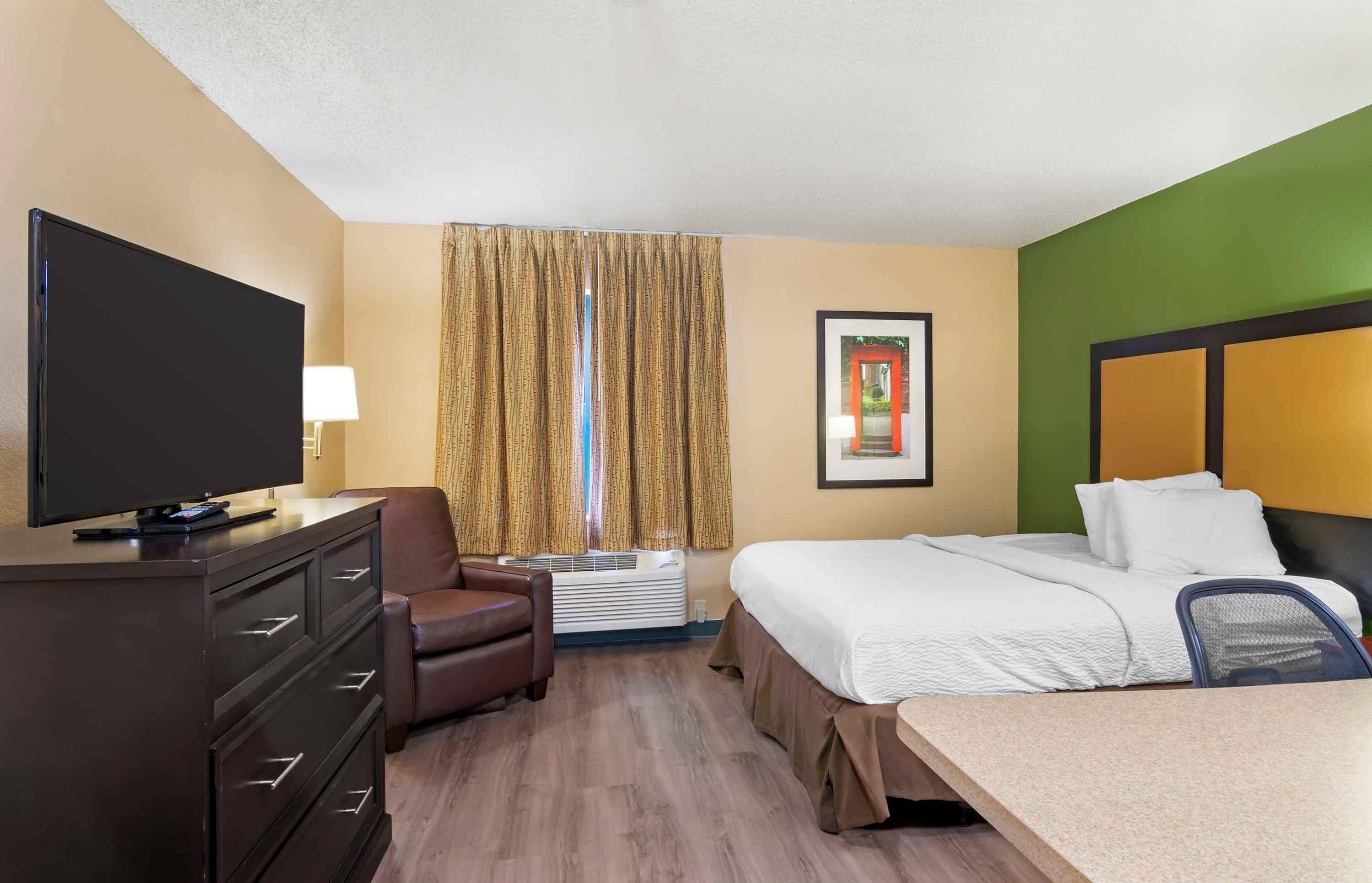 Una habitación de hotel con cama, escritorio, televisor, silla y ventana con cortinas.