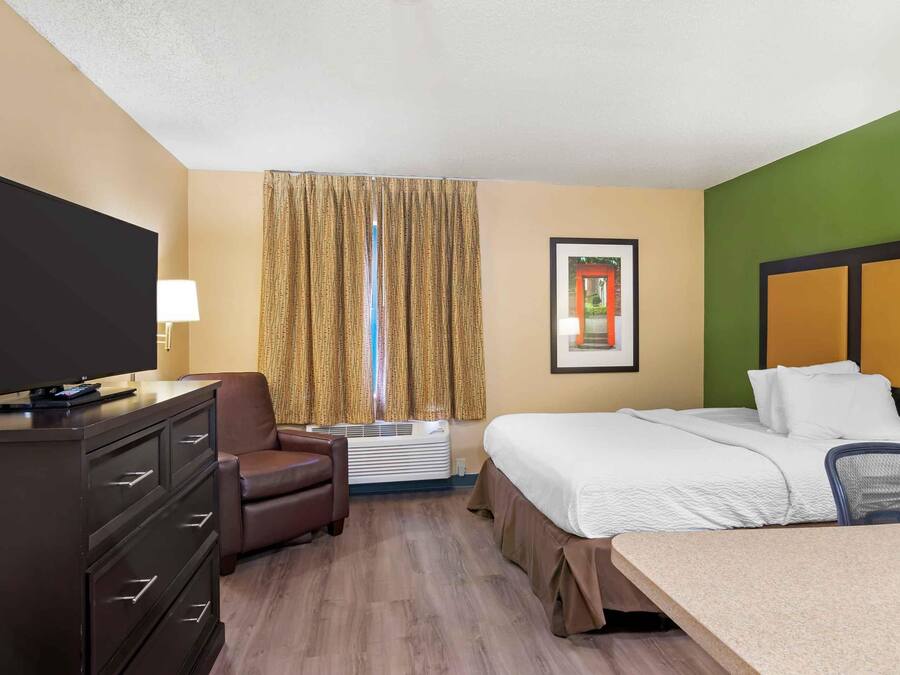 Quarto de hotel com cama, escrivaninha, televisão, cadeira e janela com cortinas.