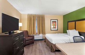 Una habitación de hotel con cama, escritorio, televisor, silla y ventana con cortinas.
