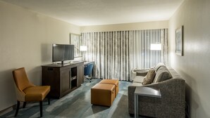 A hotel room with a flat-screen TV on a wooden desk, a brown leather chair, a gray sofa, a small ottoman, and a side table. - Hampton Inn & Suites Jackson-Coliseum (Jackson)