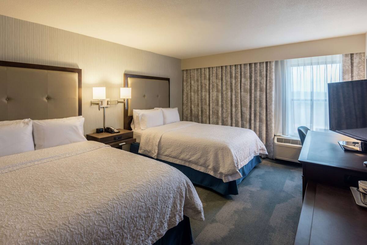 A hotel room with two beds, a desk, a chair, and a large window with curtains.