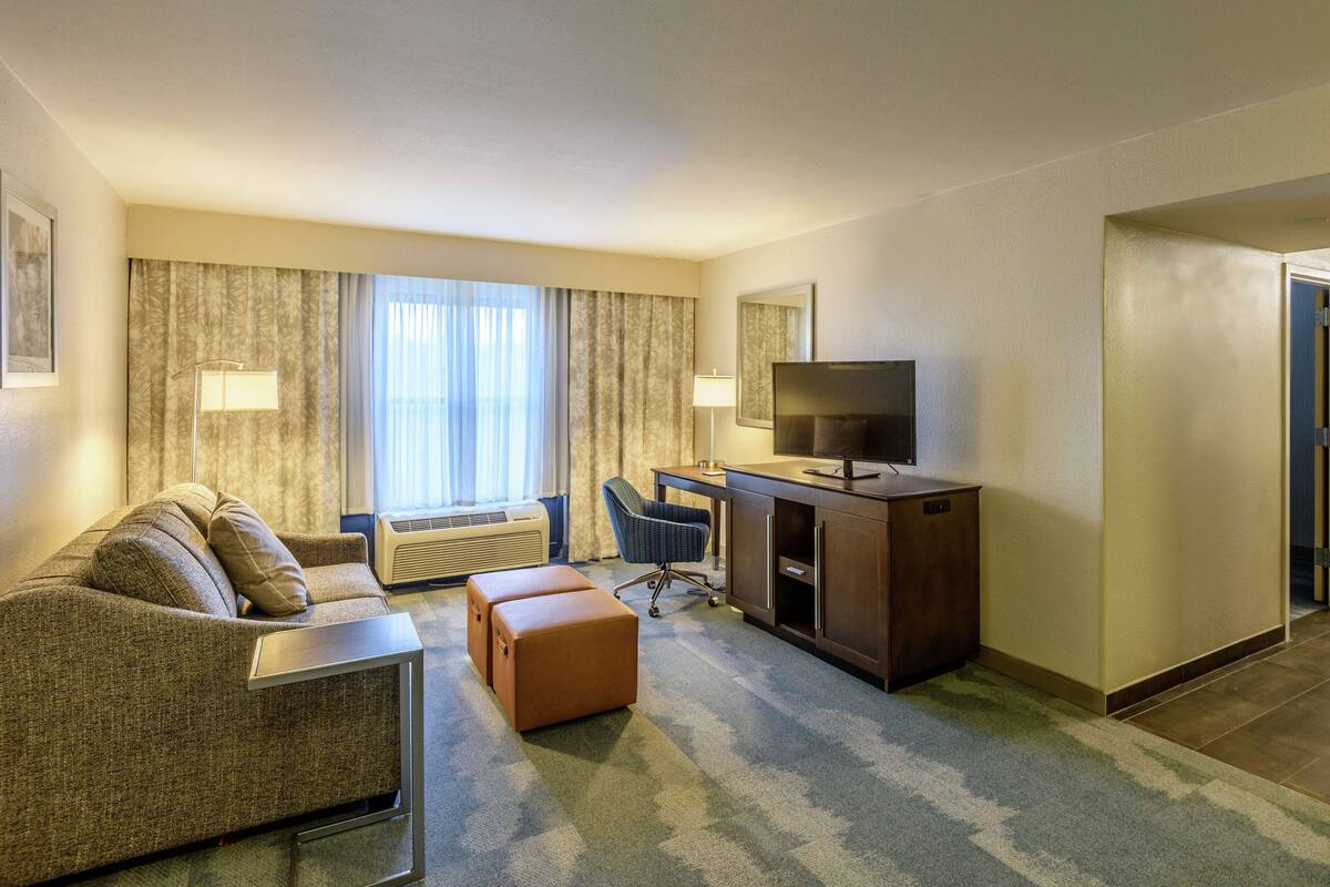 A hotel room with a sofa, a desk, a television, and a large window with curtains.
