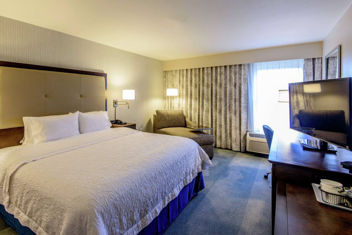 A hotel room with a large bed, a chair, a desk with a TV, and a window with curtains.
