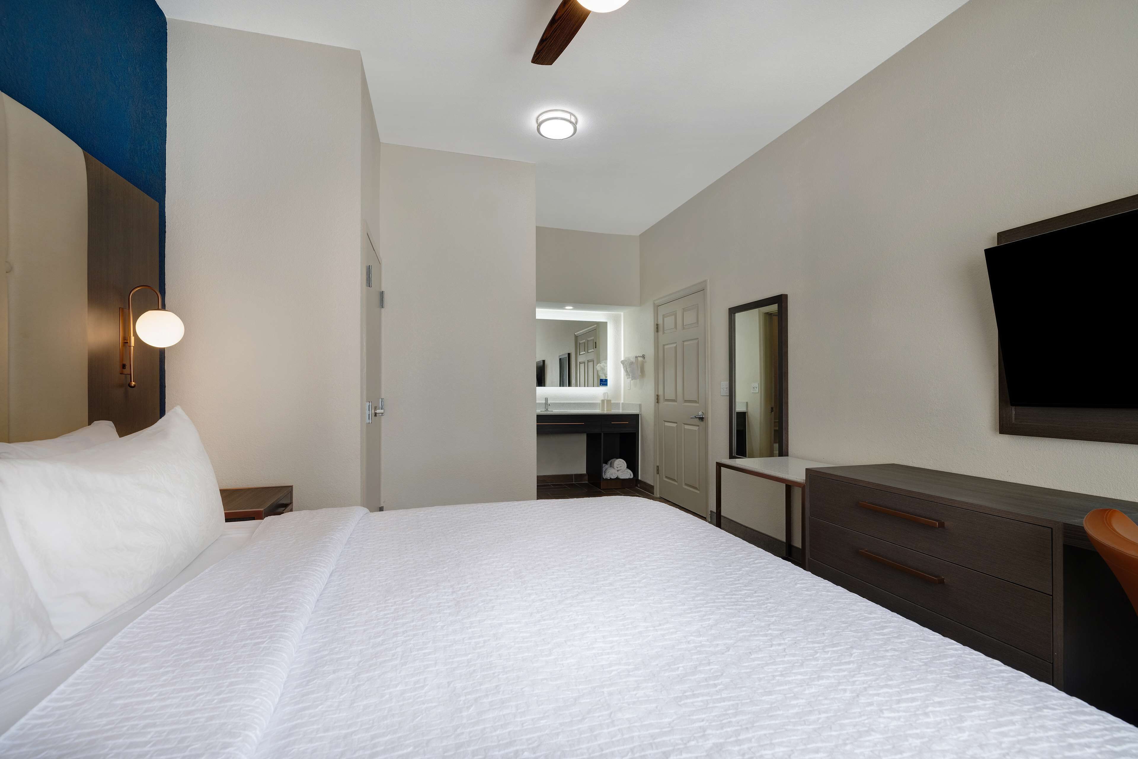 A hotel room with a large bed, a desk, a chair, a mirror, a television, and a bathroom visible through an open door.