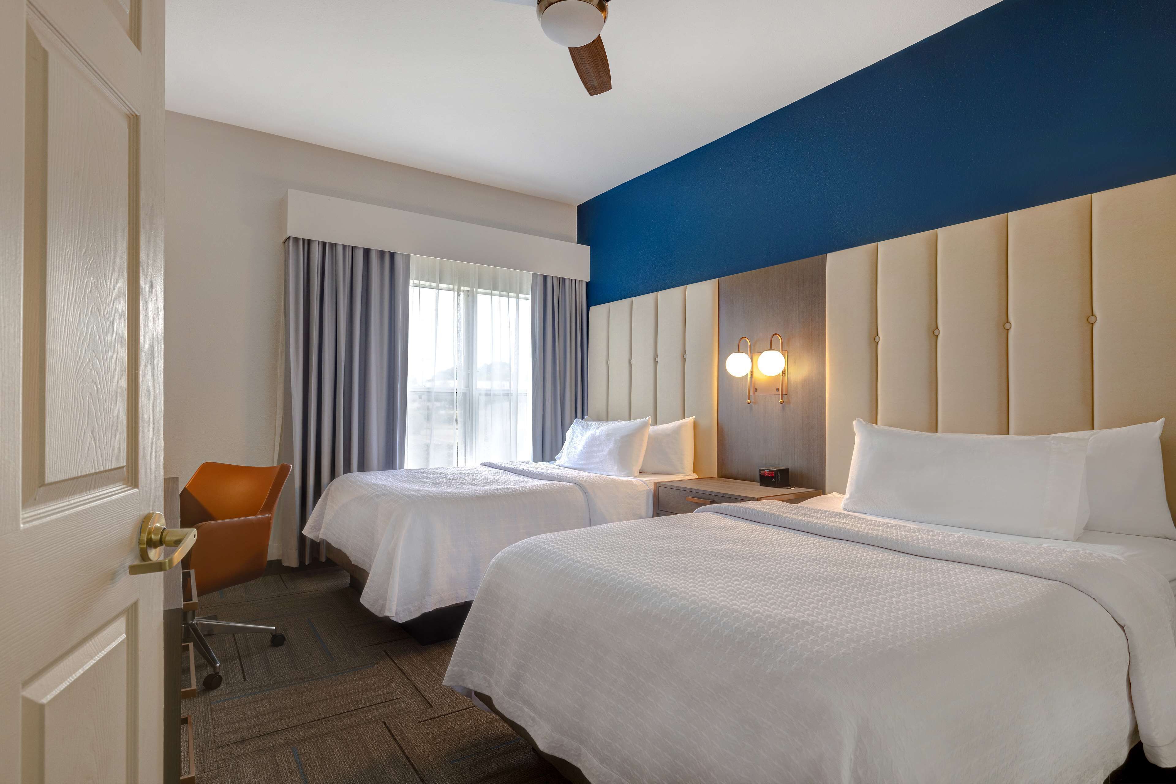 A hotel room with two beds, a desk, a chair, and a large window with curtains.