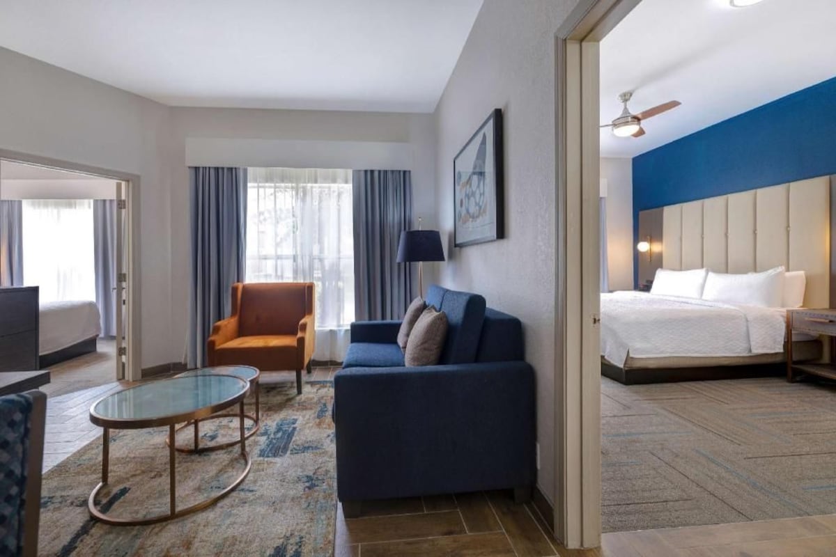 A hotel room with a bed, a sofa, a chair, a glass coffee table, and a television.