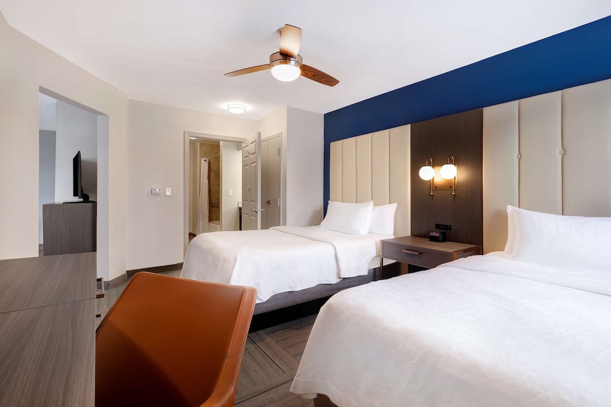 A hotel room with two beds, a desk, a chair, a ceiling fan, and a wall-mounted light fixture.