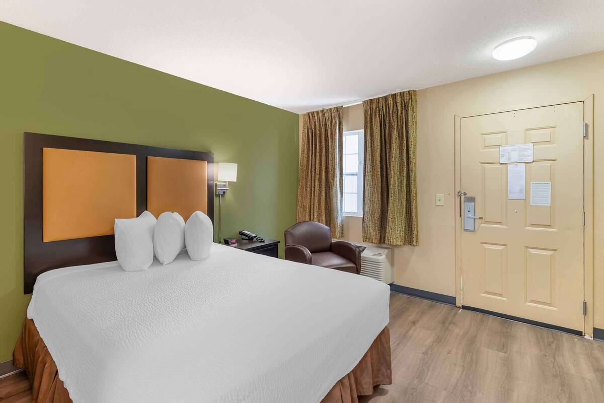 A hotel room with a large bed, a chair, a desk, and a door with a sign.