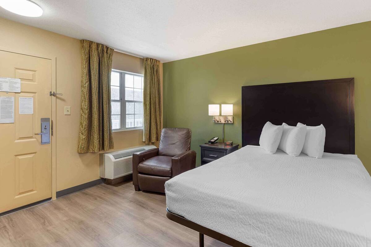 A hotel room with a bed, a chair, a nightstand, a lamp, a window with curtains, and a door with a bulletin board.