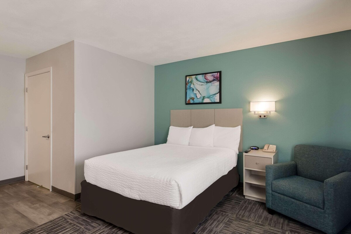 A hotel room with a bed, a chair, a nightstand, and a painting on the wall.