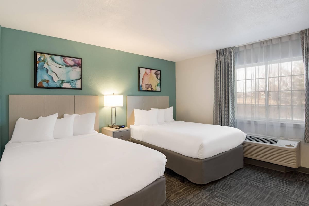 A hotel room with two beds, a nightstand with a lamp, a window with sheer curtains, and two abstract paintings on the wall.