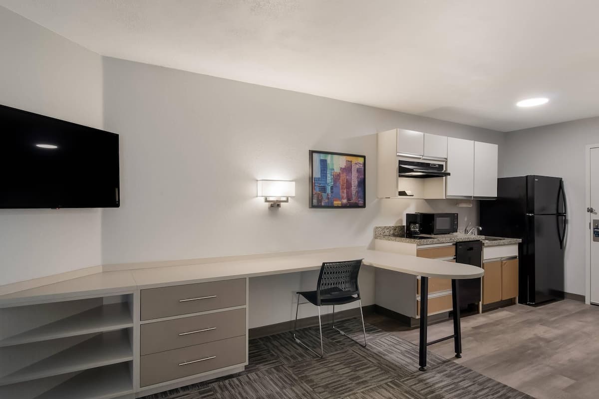 A compact kitchen with a flat-screen TV, a microwave, a refrigerator, and a small dining area with a chair.