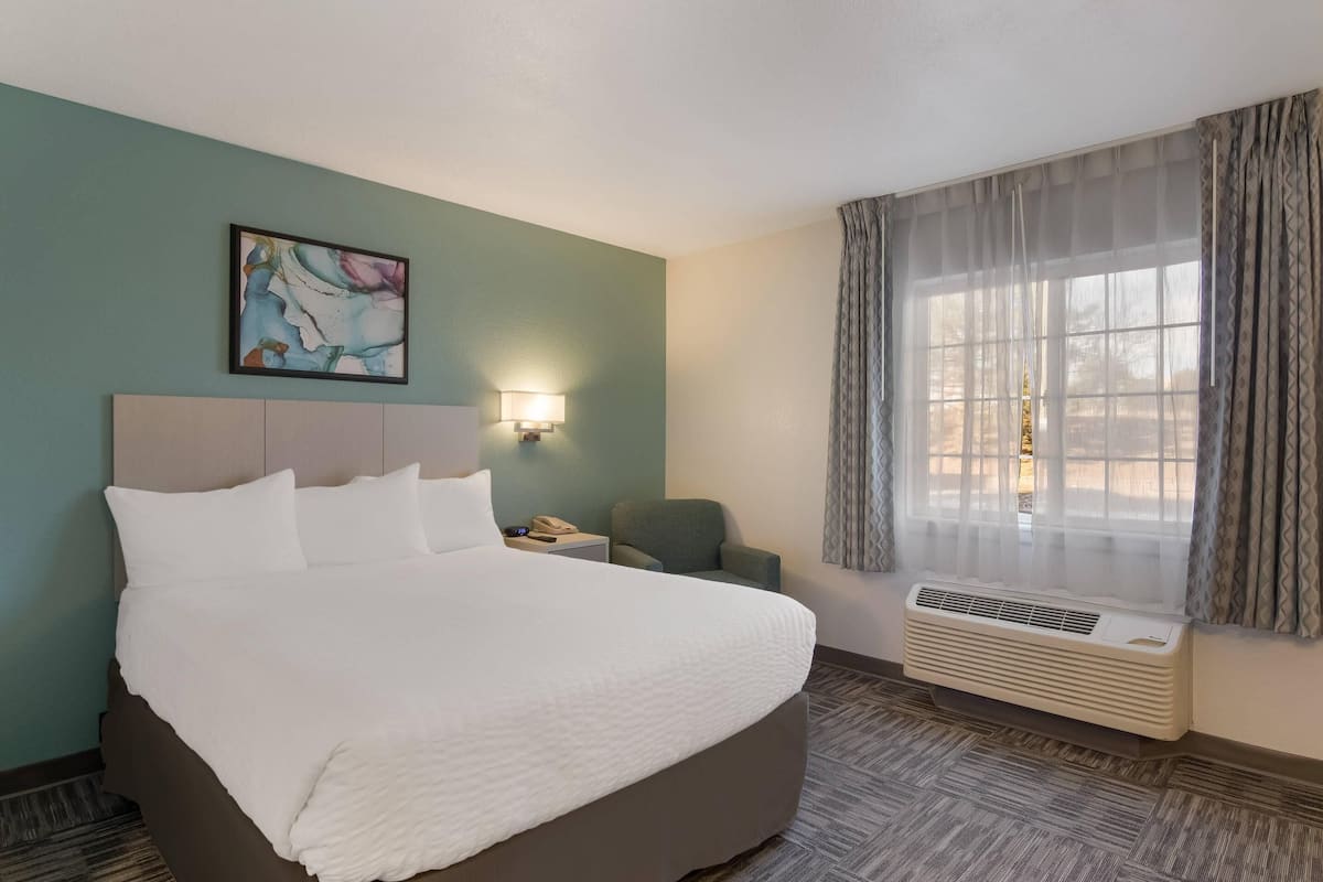 A hotel room with a large bed, a nightstand with a lamp, a chair, a window with curtains, and a wall-mounted air conditioner.