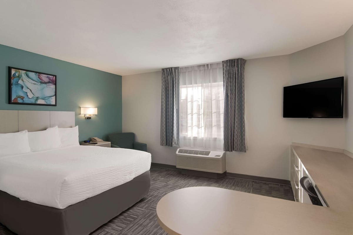 A hotel room with a large bed, a desk, a chair, a flat-screen TV, a window with curtains, an air conditioning unit, and a painting on the wall.