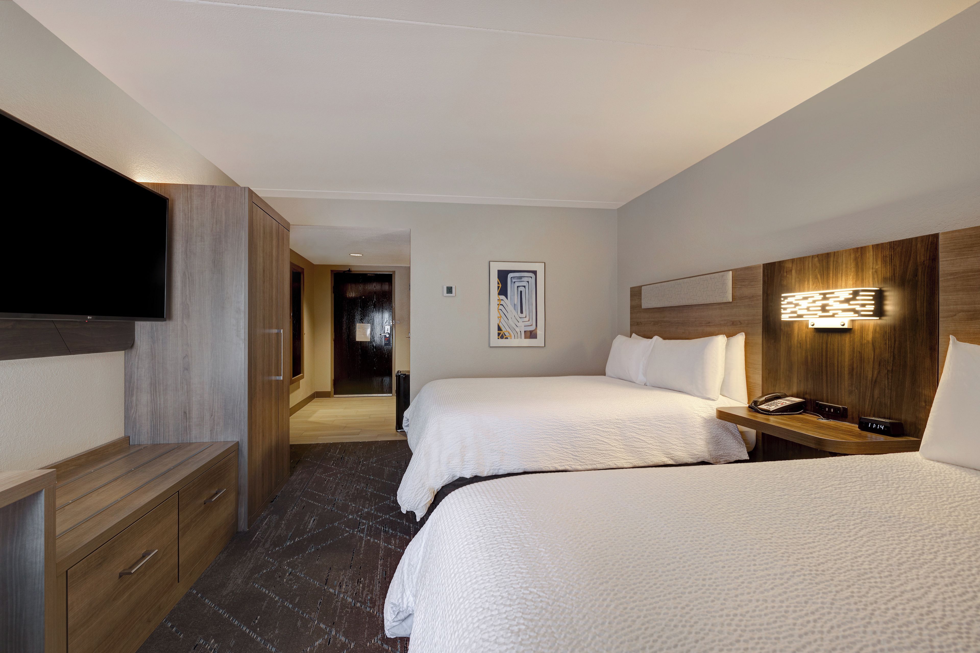A hotel room with two beds, a TV, a bedside table with a lamp, and a wooden headboard.