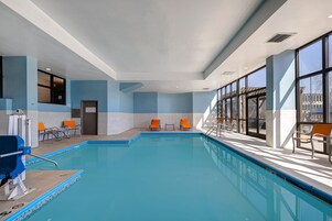 Une piscine intérieure à l’eau d’un bleu cristallin, entourée de chaises longues orange et grises, et de grandes fenêtres offrant une vue sur l’extérieur.