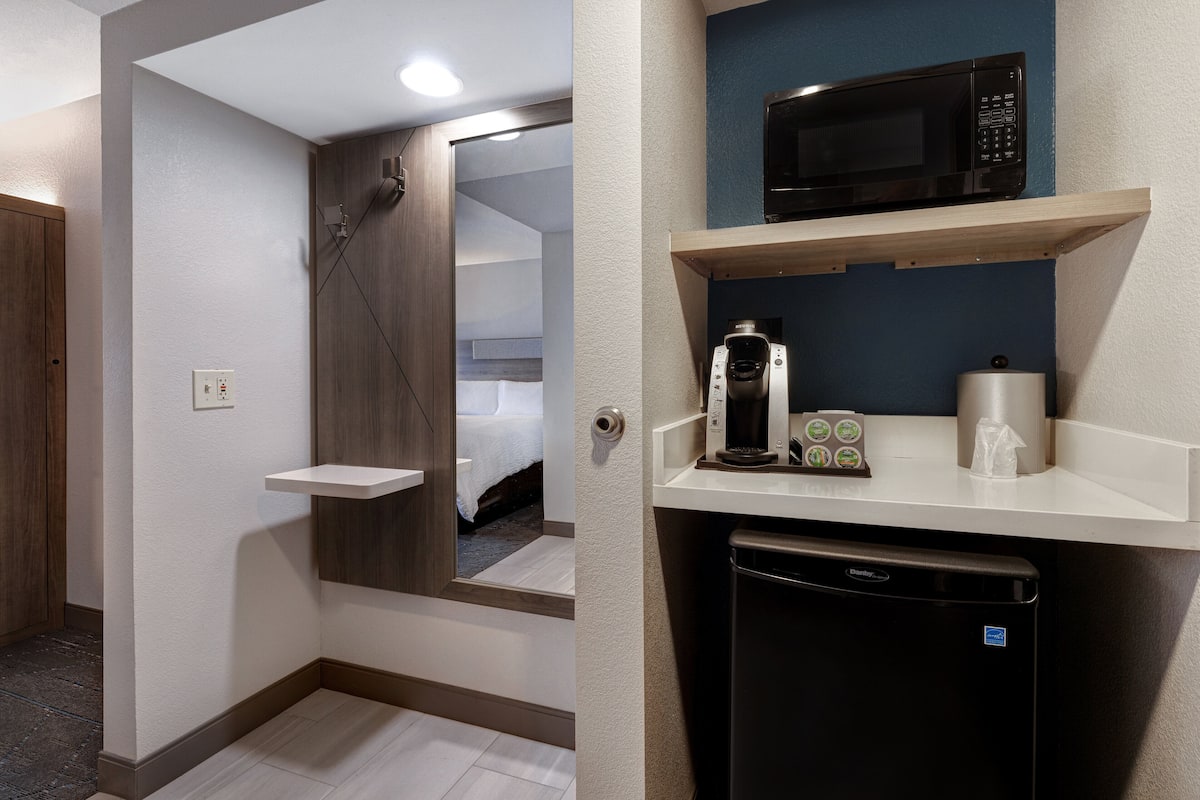A hotel room with a wooden door, a white wall, a small white shelf, a black microwave, a coffee maker, and a black dishwasher.