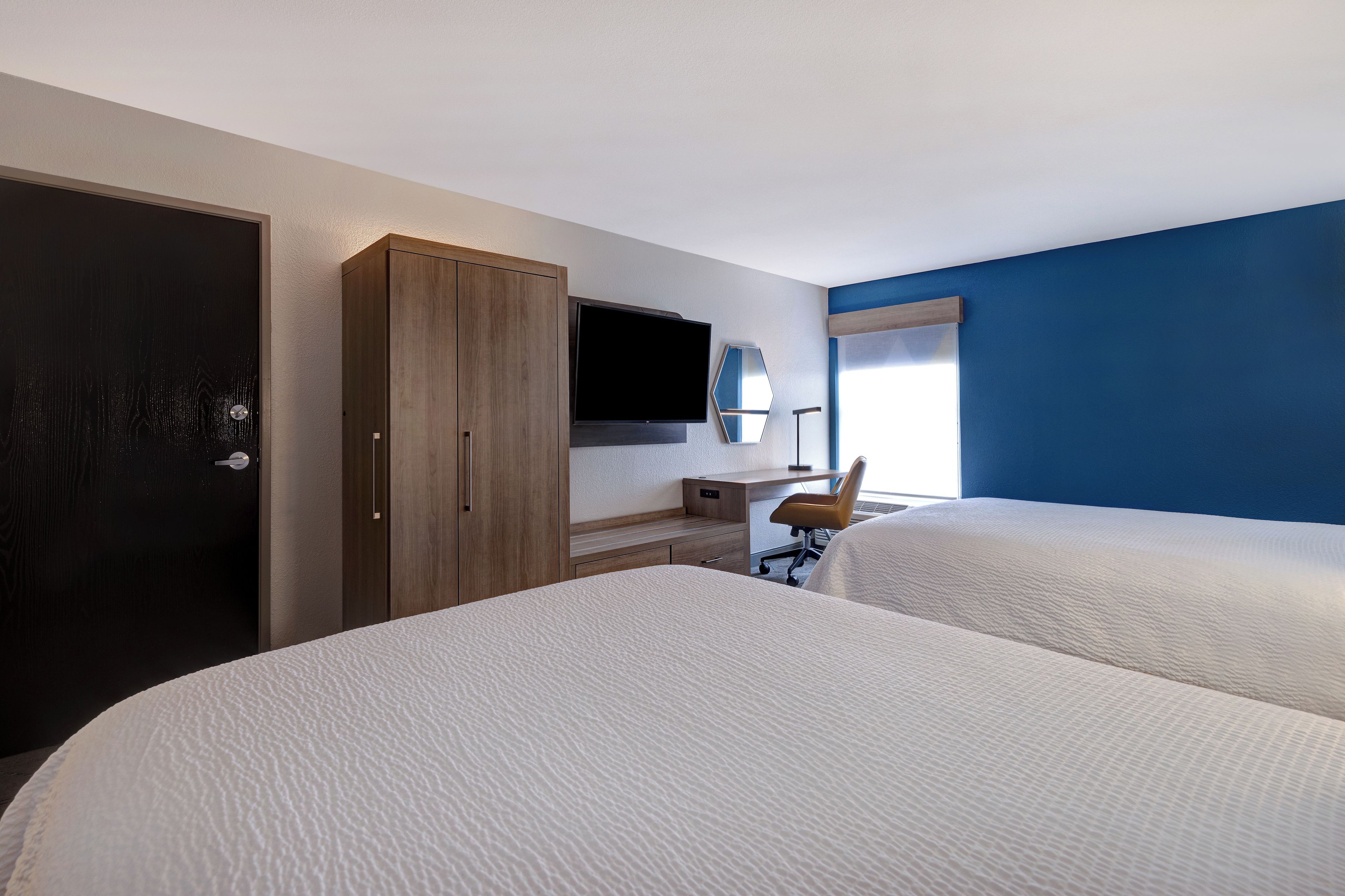 A hotel room with two beds, a desk, a chair, a wardrobe, a TV, and a window with blinds.