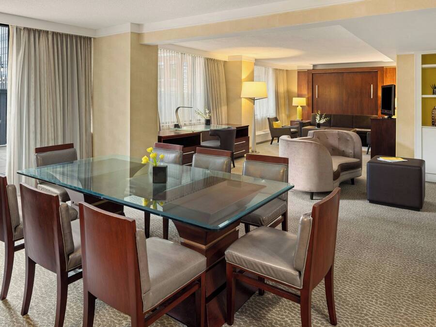 Habitación de hotel con una mesa de vidrio y sillas, un sofá, un televisor y un mueble con adornos.