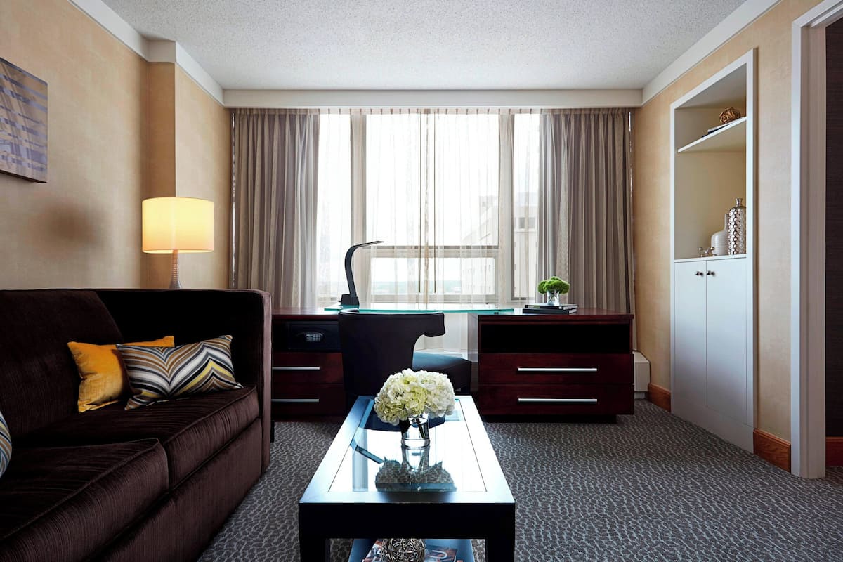 A hotel room with a dark brown sofa, a glass coffee table with a vase of flowers, a desk with a computer, and a wardrobe.