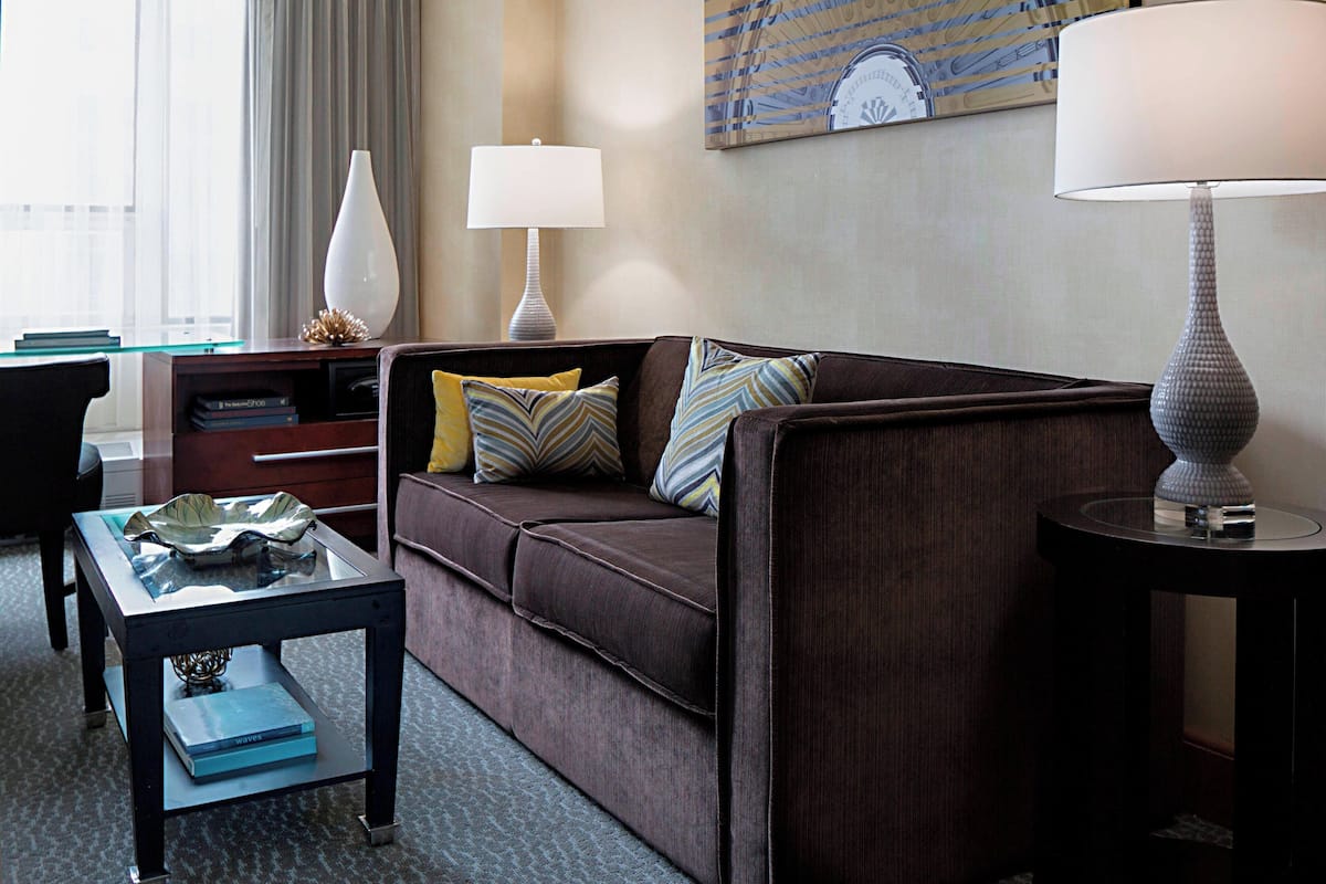 A modern hotel room with a brown sofa, a glass coffee table, a side table, and a lamp.