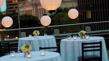 Una terraza con mesas redondas, cada una decorada con velas y centros de mesa florales, con un edificio y follaje de fondo.