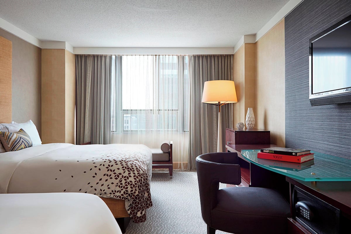 A hotel room with two beds, a desk with a lamp, a TV, and a view of the city.